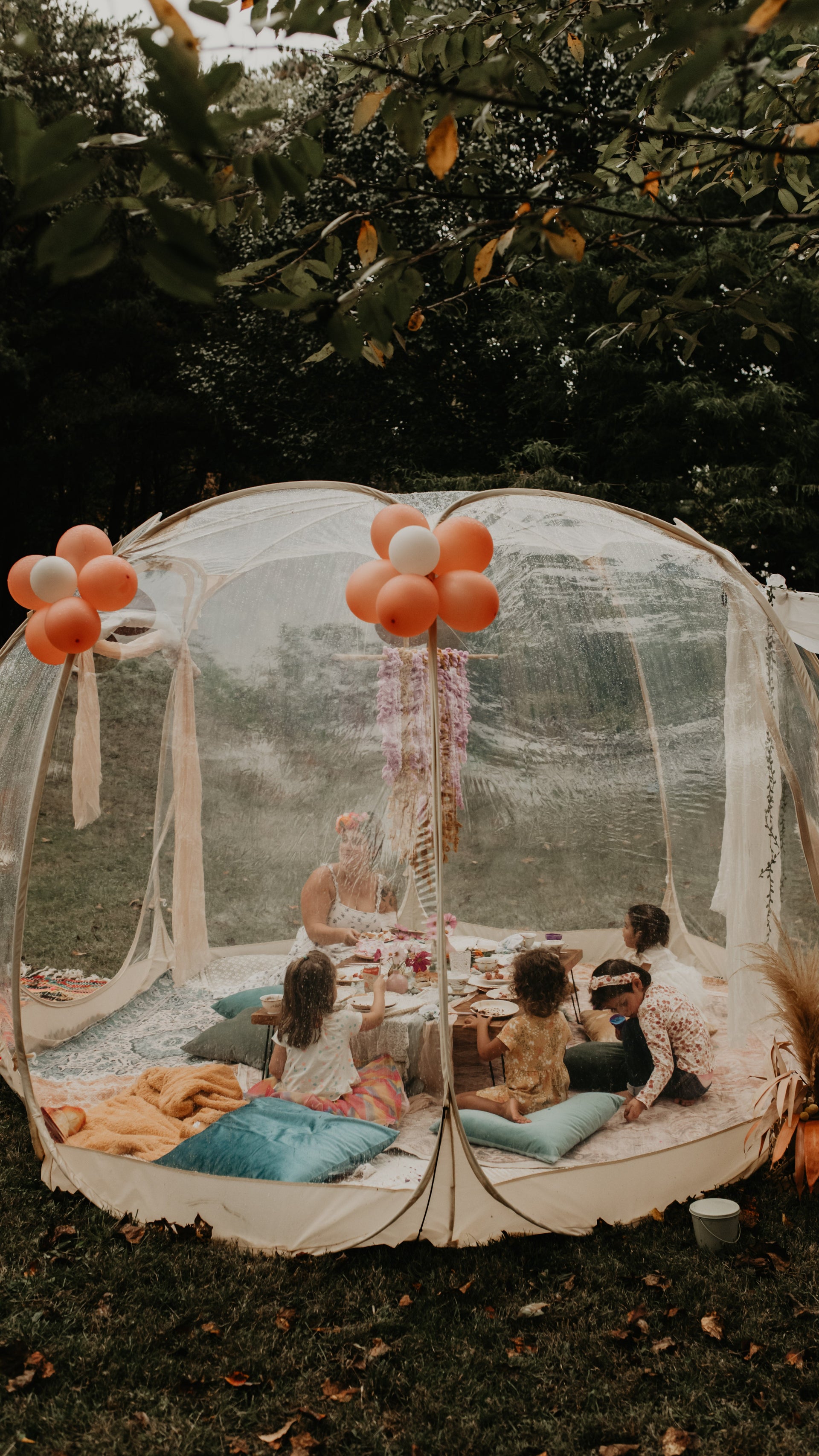 Children sitting under a transparent outdoor canopy with balloons, surrounded by greenery.  asheville luxury picnics. pop-up picnic. picnic pop-up avl