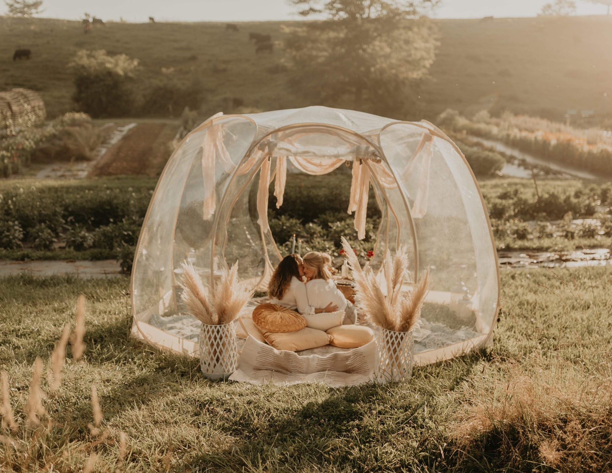 Two people sitting inside a transparent dome tent in a field with scenic background. Asheville Luxury Picnics. Picnic Pop-Up. AVL